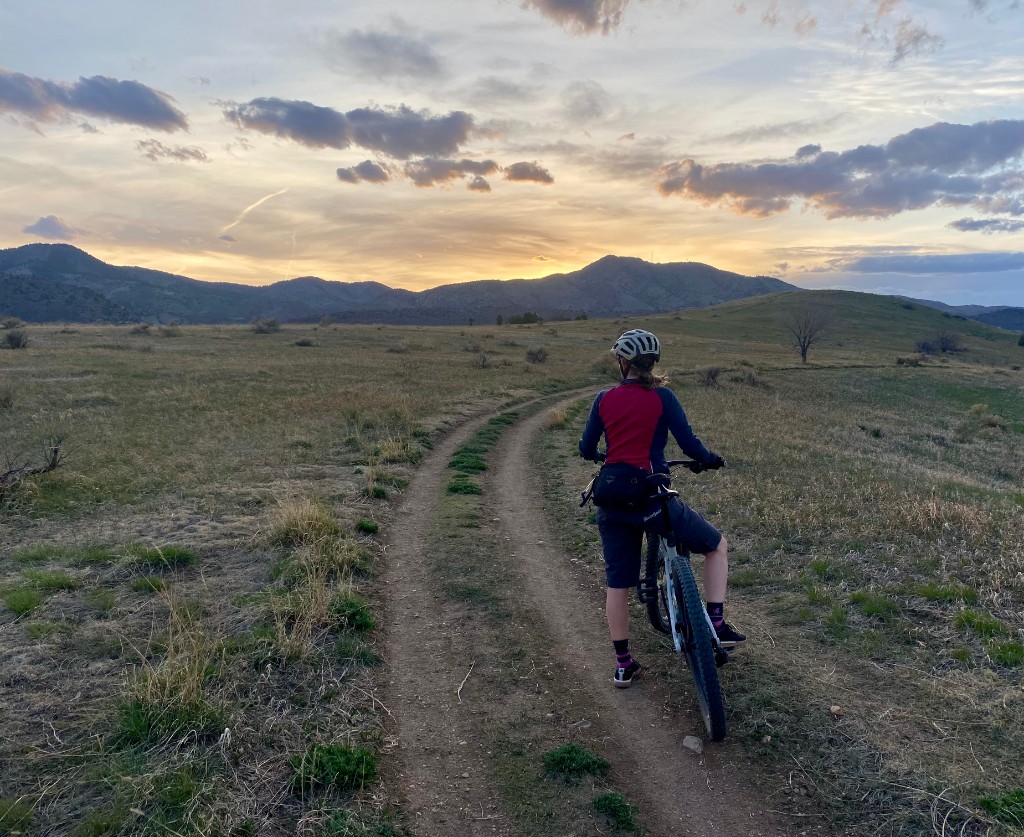 A cyclist pauses on a dirt trail at sunset, looking out over open fields toward distant mountains.
